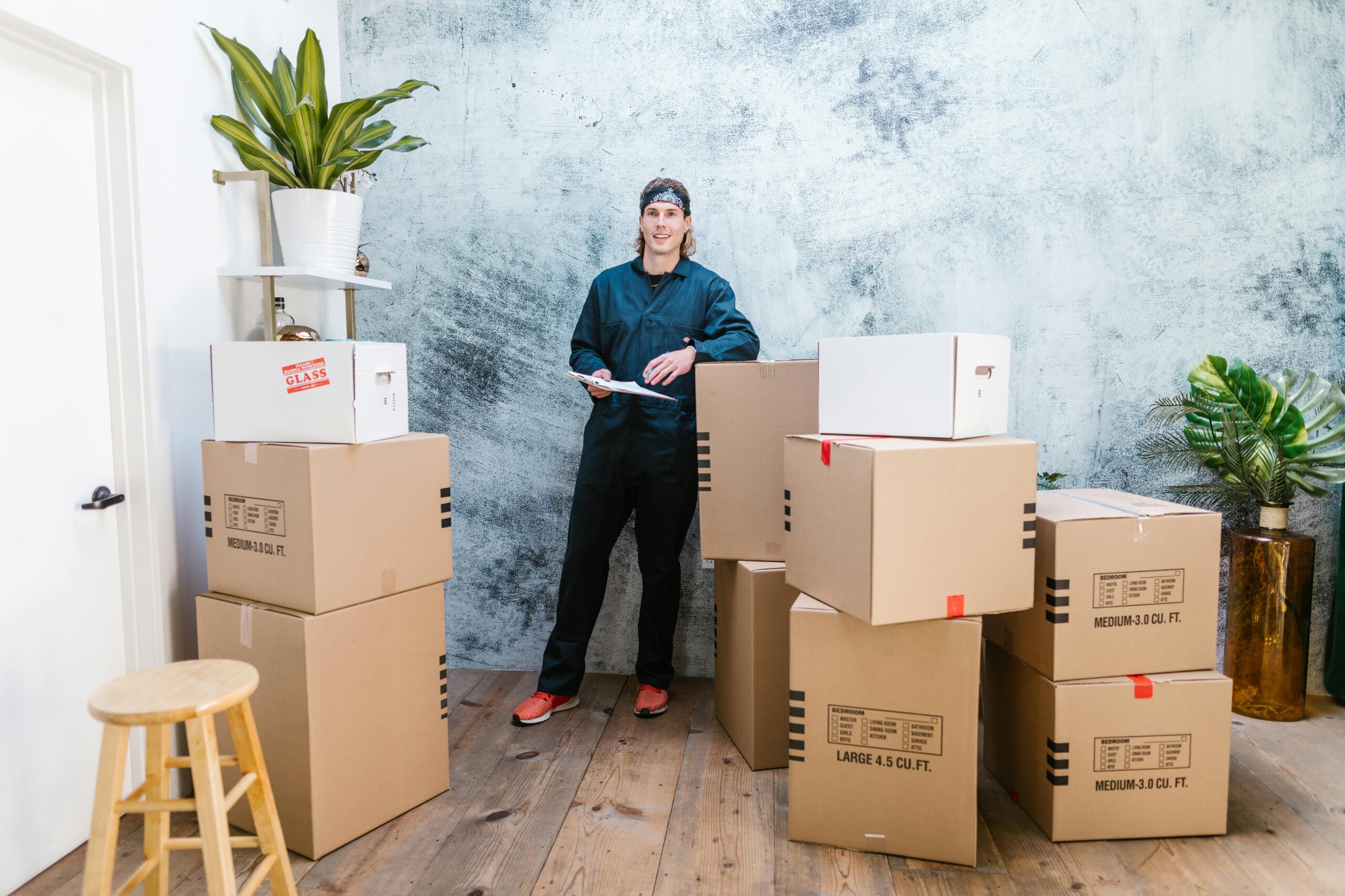Smiling mover in coveralls standing with parcels, clipboard in hand, indoors.
